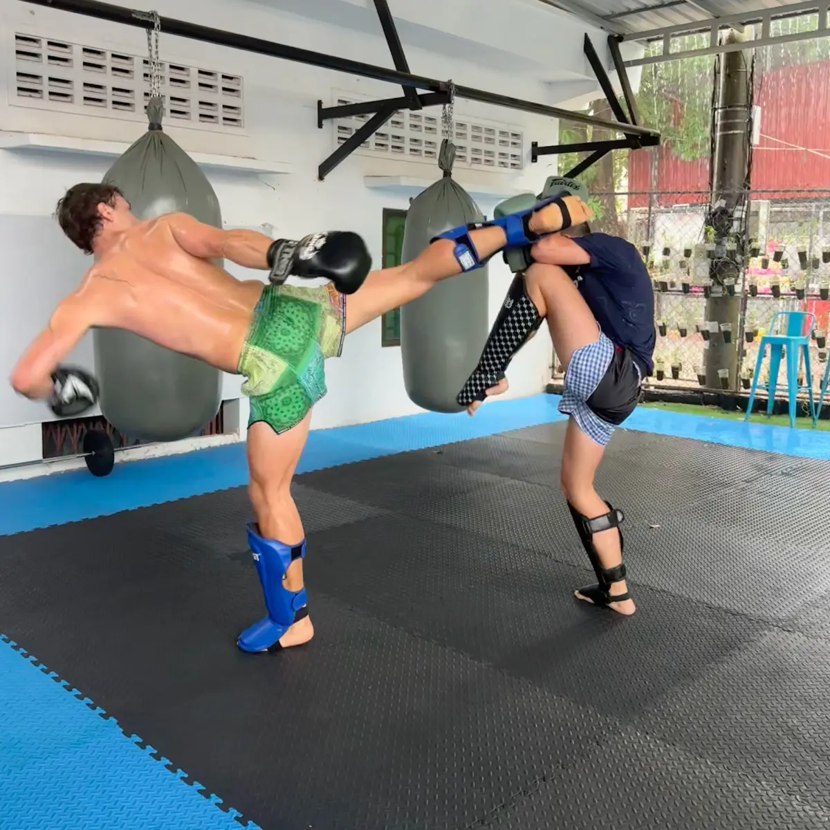 Two fighters sparring, one throwing a high kick by the heavy bags.